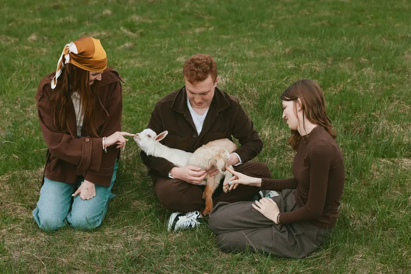 Lifestyle scene shows a family in a grassy park with a playful goat kid, three people gently interacting, feeding, and bonding, capturing everyday farm charm and curiosity, a moment of calm, care,