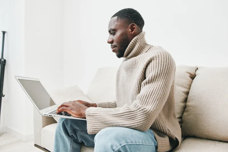 Young African man working on a laptop at home, wearing a cozy beige sweater, focused expression, modern interior setting with soft colors