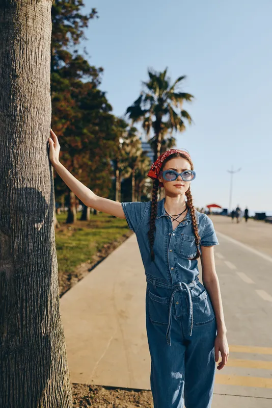 Young woman wearing sunglasses and denim jumpsuit leans on palm tree outdoors under clear sky, stylish and relaxed with braided hair and red headband in sunny setting.