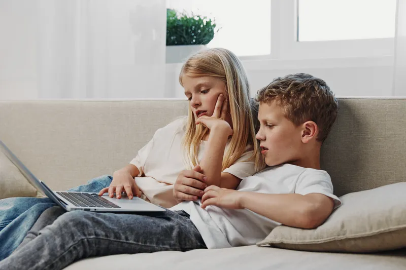 Two focused children sitting on a couch using laptop together in bright room. Concept of learning, technology, teamwork, and childhood education at home.