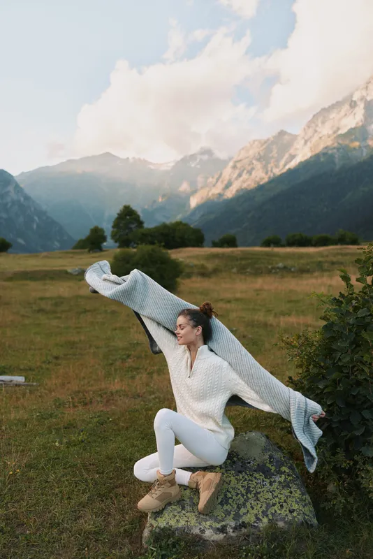 A woman in a white sweater and boots leaps across a meadow, with mountains in the background, capturing a vibrant outdoor lifestyle moment in nature.
