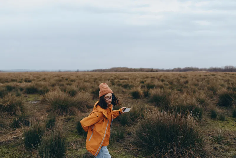 Woman lifestyle outdoors field grass, smartphone in hand, beanie and orange jacket, nature cloudy sky, she stands in the meadow, looking at screen, calm walking portrait