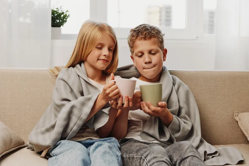 Two children wrapped in cozy blankets hold warm mugs while sitting on a sofa in a bright living room. The scene conveys comfort, relaxation, and family bonding on a cold day.
