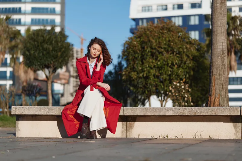 Young woman in red coat sitting on urban bench outdoors, relaxed pose, white pants, brown boots, sunny day, modern city background with trees and buildings.