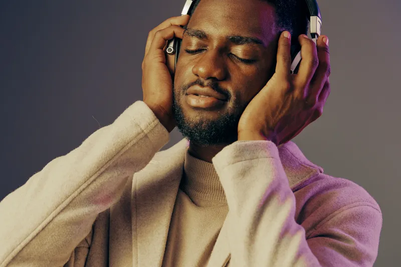 Young African man enjoying music with headphones, expressing joy and relaxation in a softly lit setting, perfect for promoting music and wellness themes
