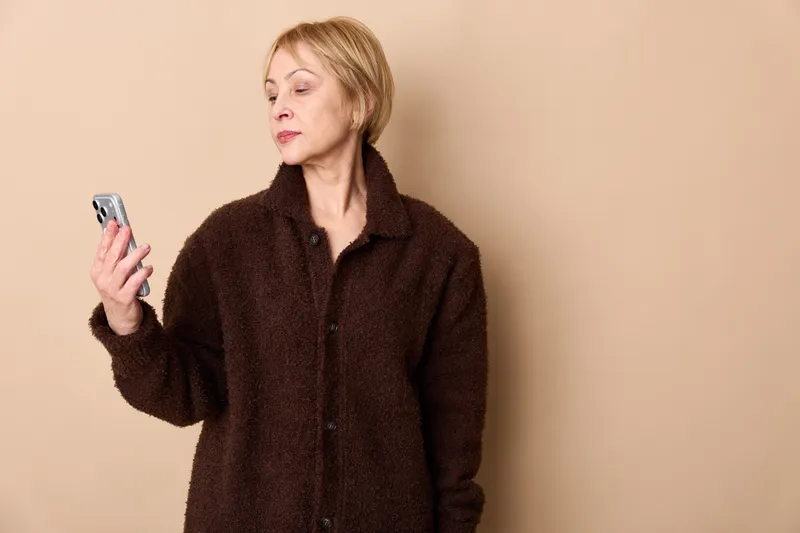 Older woman portrait holds smartphone with serious face, checking mobile messages indoors on beige studio background. Concept of communication, technology, concern.