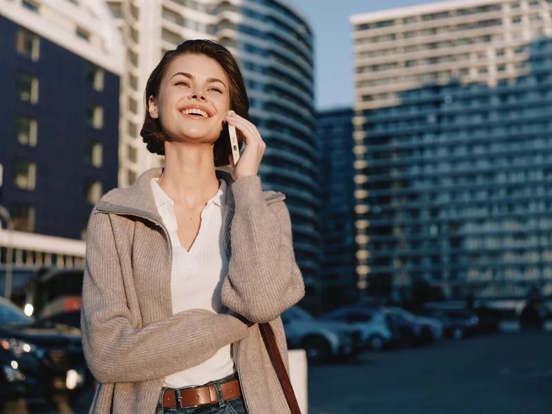 A cheerful professional woman talks on her phone in a sunlit urban setting with modern buildings, presenting a confident, relaxed vibe and a positive, productive atmosphere.