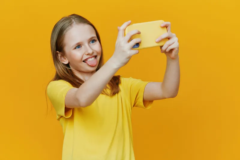 Happy girl taking a selfie with a smartphone against a bright yellow background, showcasing joy and playfulness in a casual yellow outfit