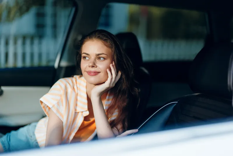 Расслабленная молодая женщина с длинными волосами отдыхает в салоне автомобиля, улыбаясь и глядя в окно