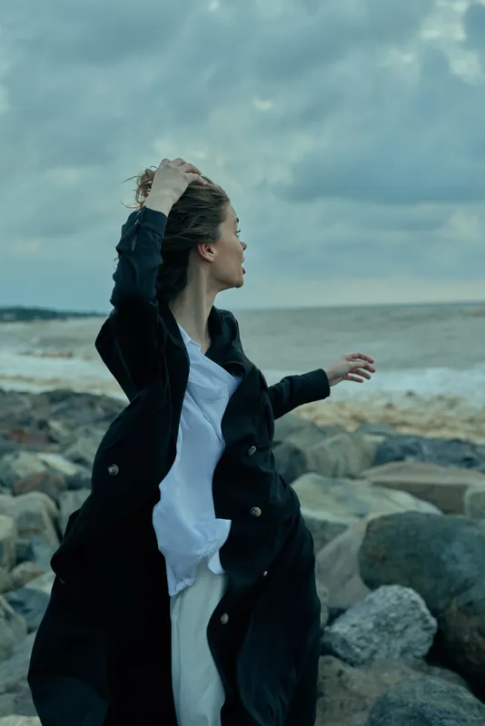 Coastline scene with windblown woman by a rocky shore, coat fluttering, ocean horizon and moody sky, motion and style, outdoor fashion mood, dramatic sea breeze and nature inspired storytelling