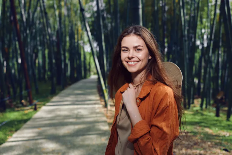 Woman smile in forest on a paved path, backpack and hat on shoulder, nature portrait of a happy traveler enjoying an outdoor walk with sunlight filtering through trees
