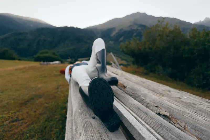 Person lying on wooden beam with boots in a meadow, mountains in background. Relax and nature scene with casual clothing, outdoor leisure, countryside landscape and peaceful rest.