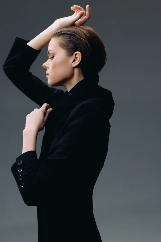 stylish woman in black blazer posing sideways with closed eyes and serene expression against gray background