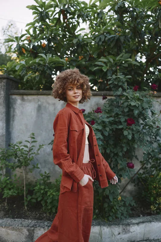 Young woman with curly hair wears a rust suit in a garden, posing confidently as soft sunlight filters through lush greenery behind her