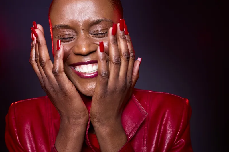 Close-up of a happy dark skinned African woman with short hair wearing red jacket and red nail polish smiling with closed eyes and hands on face. Concept of joy and beauty.