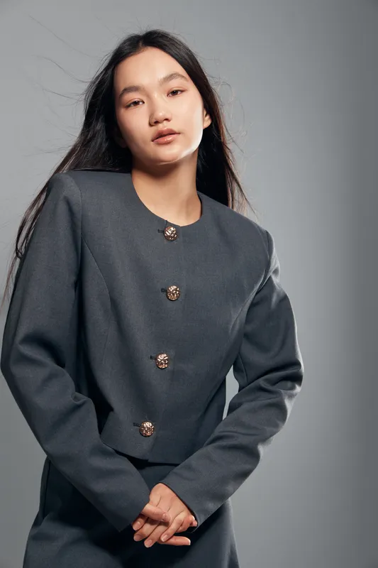 Woman with long hair wearing dark gray blazer standing with hands clasped in front against neutral background for fashion portrait, style, confidence and elegance.