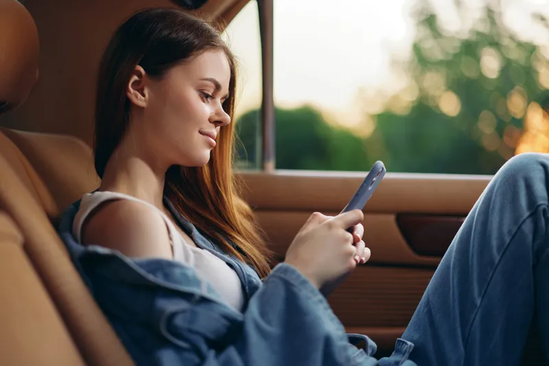 Young woman in casual denim attire relaxing in a car while using her smartphone, featuring a serene expression and a soft, natural background during sunset.