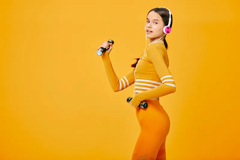 Female fitness athlete in orange athletic wear lifting dumbbells against a bold orange backdrop, showcasing dynamic movement, energy and balance in a studio workout scene.