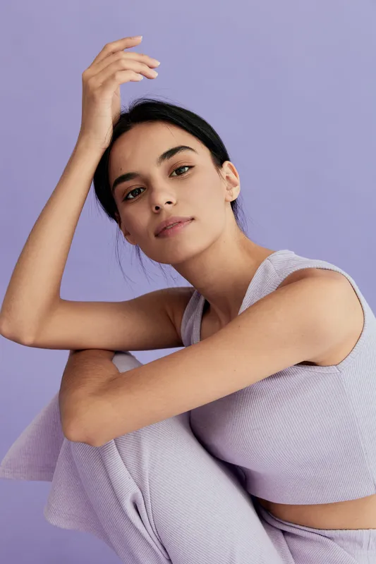 Young woman with dark hair wearing lilac knitted top and pants poses thoughtfully against a soft purple background in a minimalist studio setting.