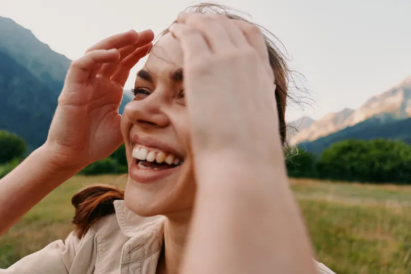 Woman smiling outdoors in mountains, enjoying sunlight and nature, a joyful lifestyle moment in a casual look, capturing a fresh, relaxed outdoor scene