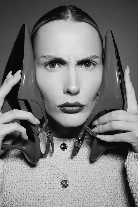 Portrait of a serious Caucasian woman with pale skin holding two high heel shoes near her face, dressed in a knitted buttoned sweater. Concept of fashion, style, and attitude.