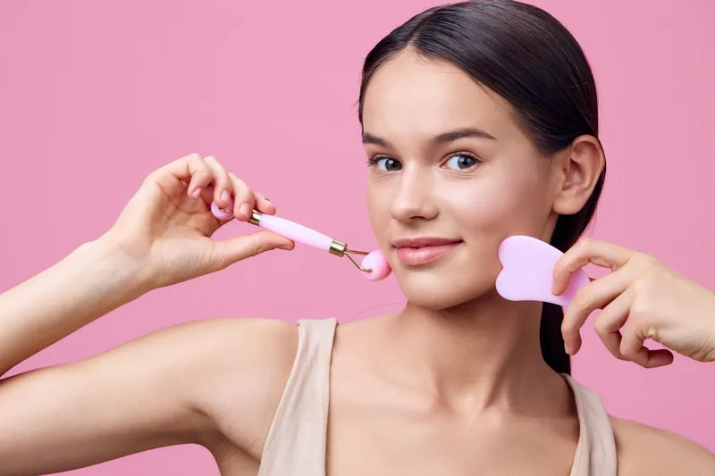 Young woman with a facial roller and heart shaped massager against a pink background, conveying beauty and self care, perfect for wellness themes