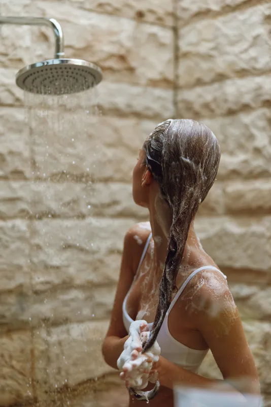 woman washing hair under shower with white soap foam, natural skin, wet brown hair, textured beige stone wall background, relaxed morning routine