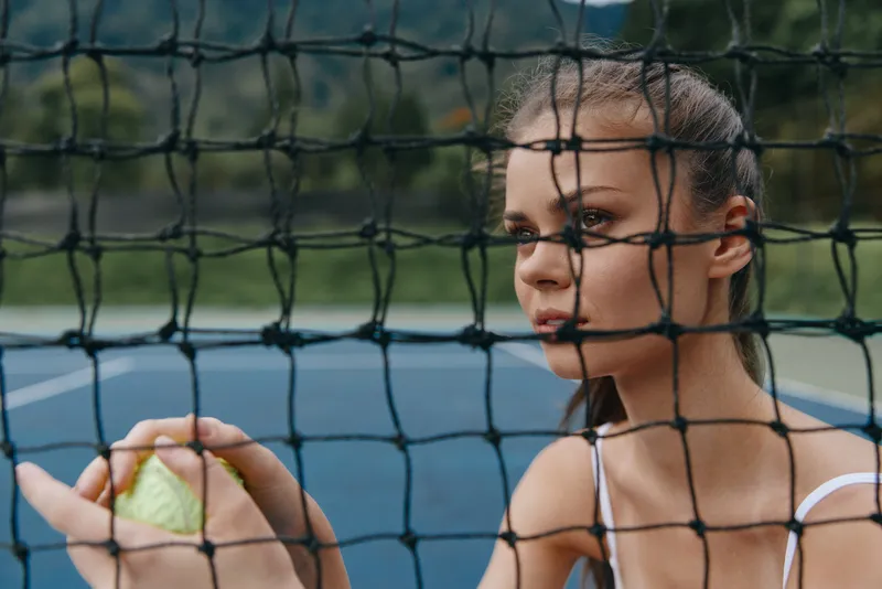 Young female tennis player holding a ball, gazing thoughtfully through the net, encapsulating determination and focus with a blurred outdoor court background