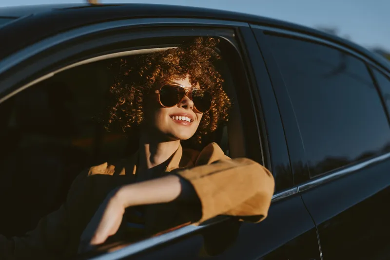 A stylish young woman with curly hair sits in a car, wearing sunglasses and a tan jacket, smiling as warm retro light pours through the window.