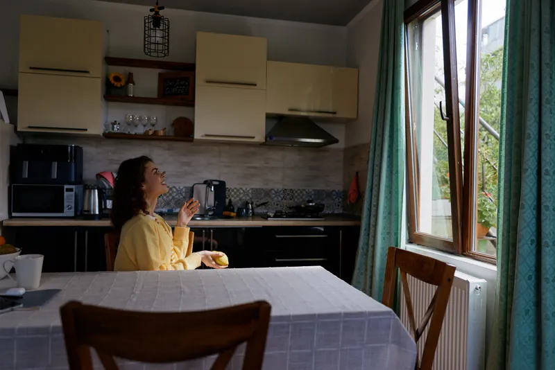 Smiling woman in a yellow top sits in a cozy kitchen, gazing toward the window. A calm domestic scene highlighting home life, selfcare, and a relaxed morning mood.
