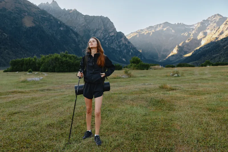 A lone hiker stands in a vast meadow beneath towering peaks, gripping trekking poles. She wears a dark jacket and shorts, exuding confidence as the wild landscape invites exploration.