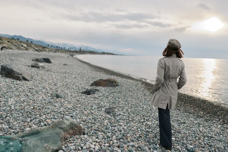 Woman in coat and hat walks alone on rocky beach during cloudy sunset. Concept of solitude, reflection, and peaceful nature near calm sea and distant mountains.