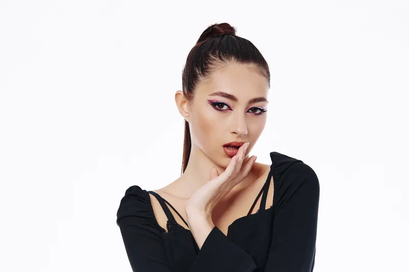 Young woman with high ponytail and striking pink eyeshadow poses with hand near mouth. Studio shot on white background. Creative concept for beauty, fashion, and editorial.