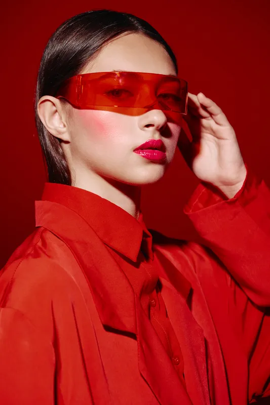 Young woman with glowing skin wearing translucent red visor and matching bold red outfit over red background. Editorial beauty concept with futuristic fashion and high gloss makeup style.
