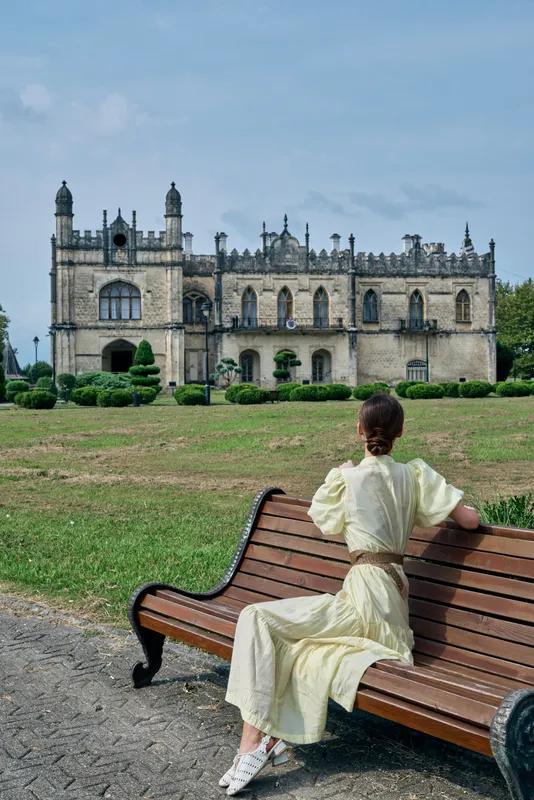 A woman in a light dress sits on a wooden bench near a wide lawn, gazing at a grand historic castle in a sunny park, creating a serene contemplative landscape