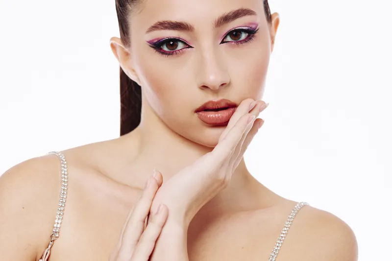 Young woman with creative makeup featuring purple eyeliner and glossy lips poses with hands near face against white background. Concept of beauty, fashion, and unique style.