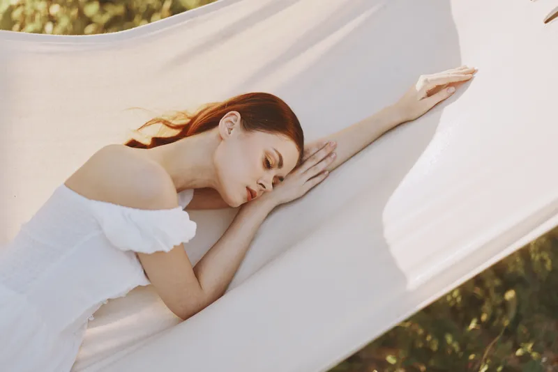 Young woman resting peacefully in a hammock, wearing a white off shoulder dress, surrounded by a serene natural backdrop, evoking calmness and relaxation