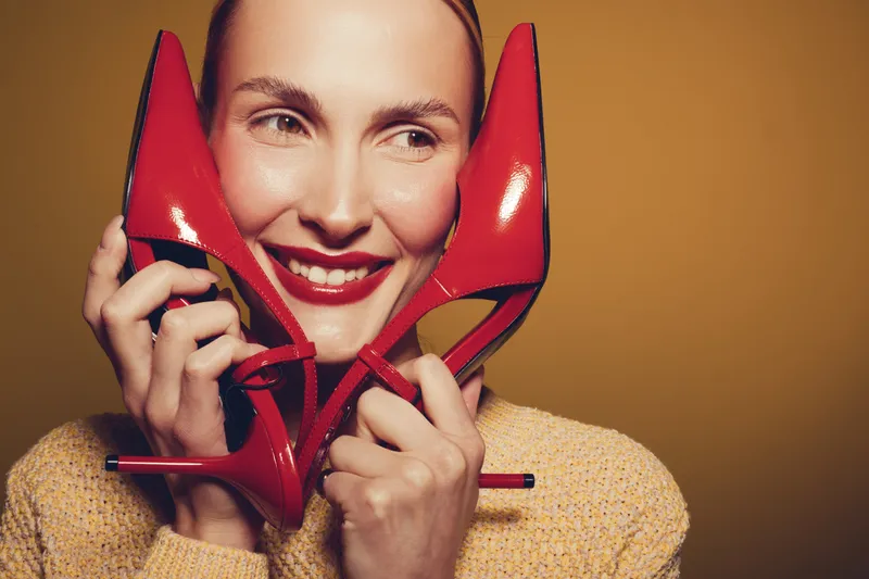 Smiling woman holds red high heels near face, wearing beige sweater. Close-up portrait on yellow background. Concept of fashion, style, and happiness with bright colors.