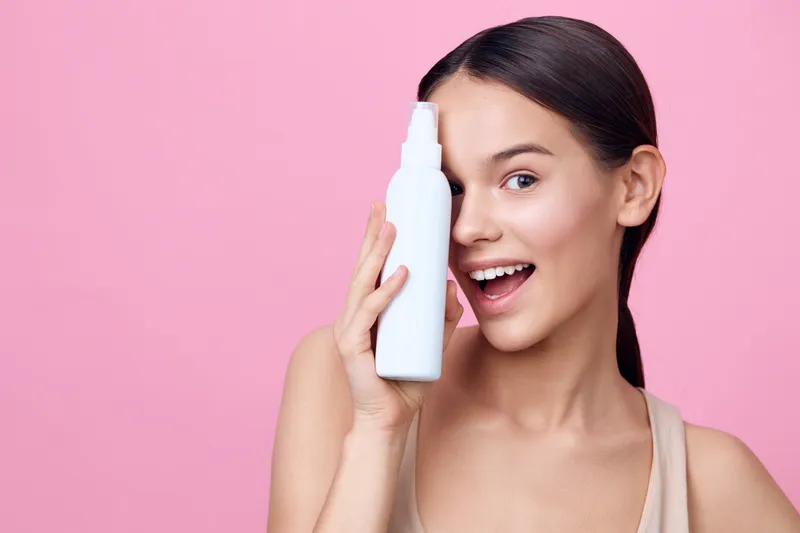 Young woman holding a white spray bottle, smiling playfully against a soft pink background, showcasing skincare and beauty concepts