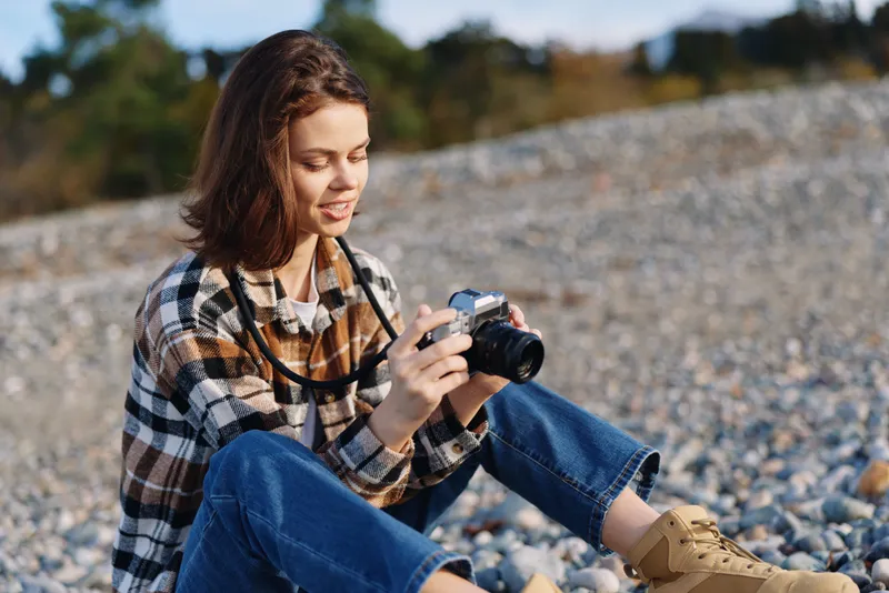 Woman photographer sits on rocky ground concentrating on her camera, capturing outdoor scenery with focus and calm, wearing plaid shirt and boots in a natural landscape.
