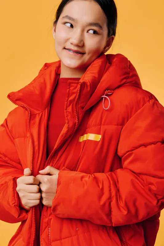 Young woman in red puffer jacket looks sideways with shy smile studio portrait, casual winter clothing fashion, cozy warm coat and soft light on yellow background.