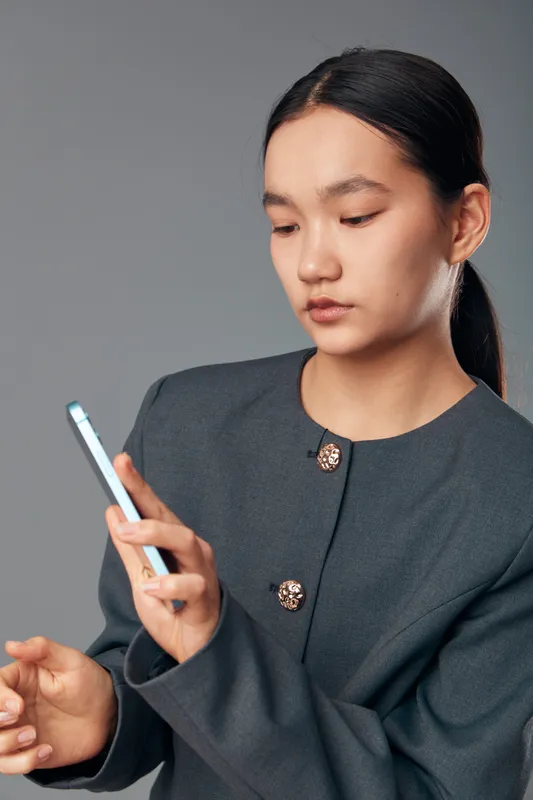 Woman with smartphone in dark professional jacket using mobile device for communication or work. Portrait of focused Asian female holding phone on gray background for technology and connection.