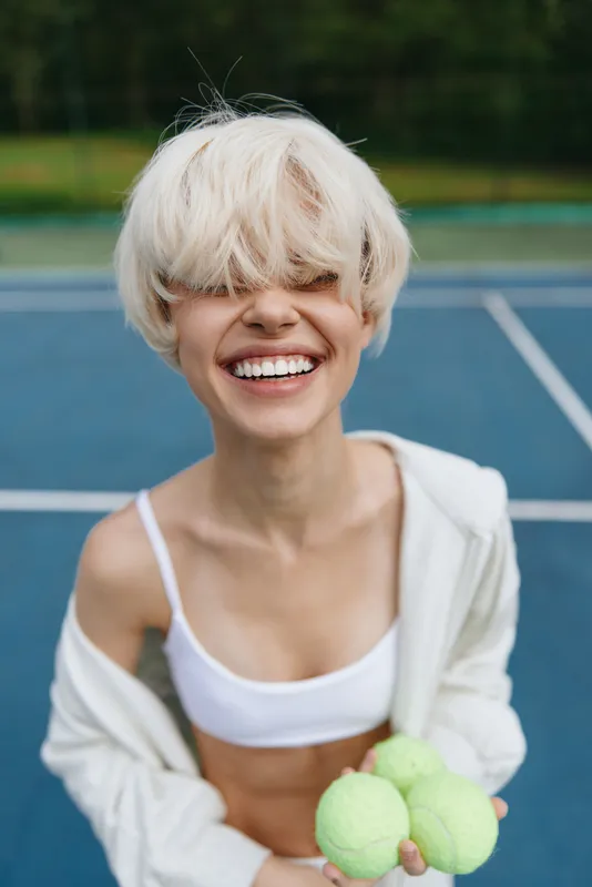 Cheerful young woman with short blonde hair holding tennis balls on a blue court, showcasing joy and an active lifestyle during summer sports activities