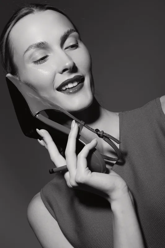 Happy Caucasian woman with light skin holds shoe as phone, smiling and looking amused. Creative black and white portrait with playful mood and stylish modern concept.