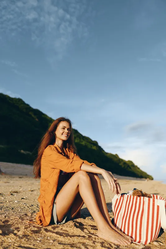 Young woman in orange shirt sitting on sandy beach with striped bag, smiling and enjoying sunny day near green hills under blue sky, concept of summer vacation and relaxation.