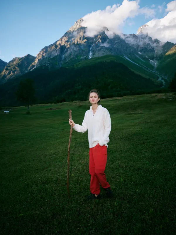 Person standing in a lush field with a wooden staff, majestic mountains in the background, white shirt and red pants, outdoor landscape conveying adventure and exploration