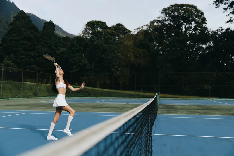 Young athletic woman in a white tennis outfit playing on a blue outdoor court, showcasing focus and determination while serving