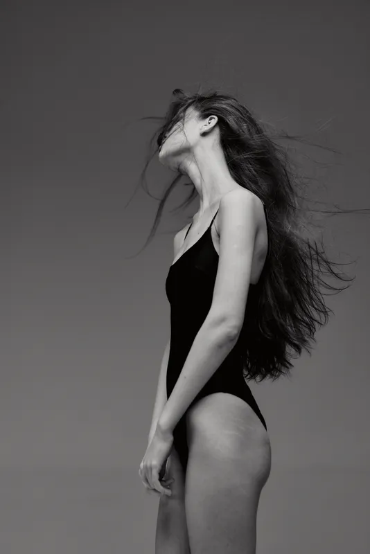Black and white portrait of a woman with long hair in a swimsuit standing against a plain background.