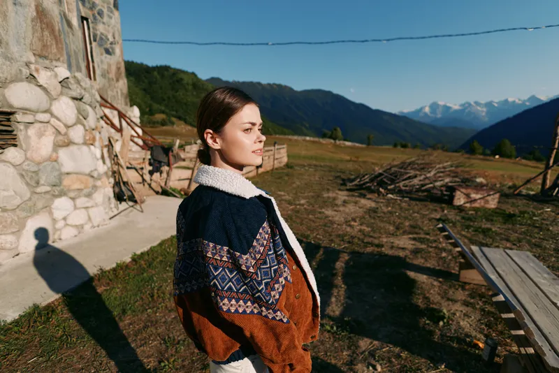 Woman portrait in countryside wearing patterned sweater near stone cottage, looking toward distant mountains at golden hour. Outdoor lifestyle, travel, nature and peaceful rural scene.
