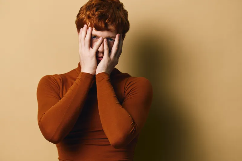 Redhead man in brown turtleneck covering face with hands looking through fingers, portrait on beige background with shadow for emotion and expression concept.